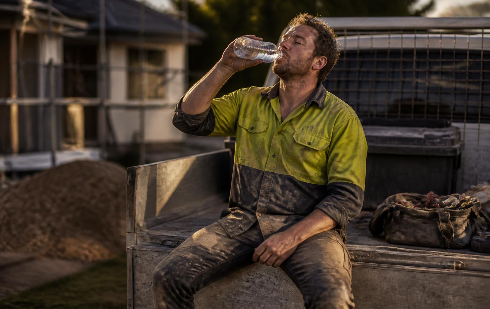 Australian worker in dusty workwear sitting on the tailgate of a ute at the end of a shift, drinking water in warm late-afternoon light