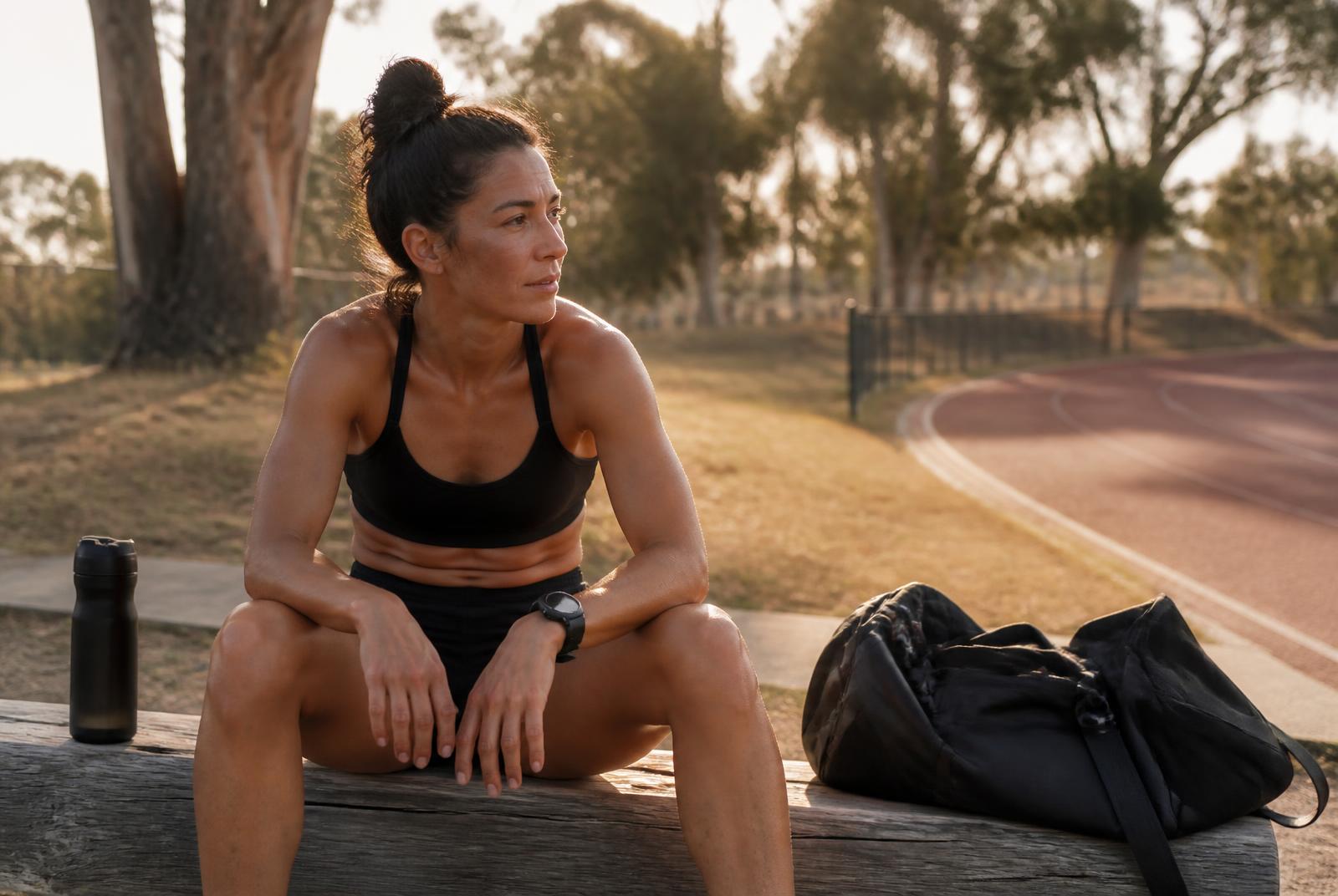 Recreational athlete sitting on a timber bench trackside beside a suburban Australian athletics track in late afternoon light, resting between training sessions
