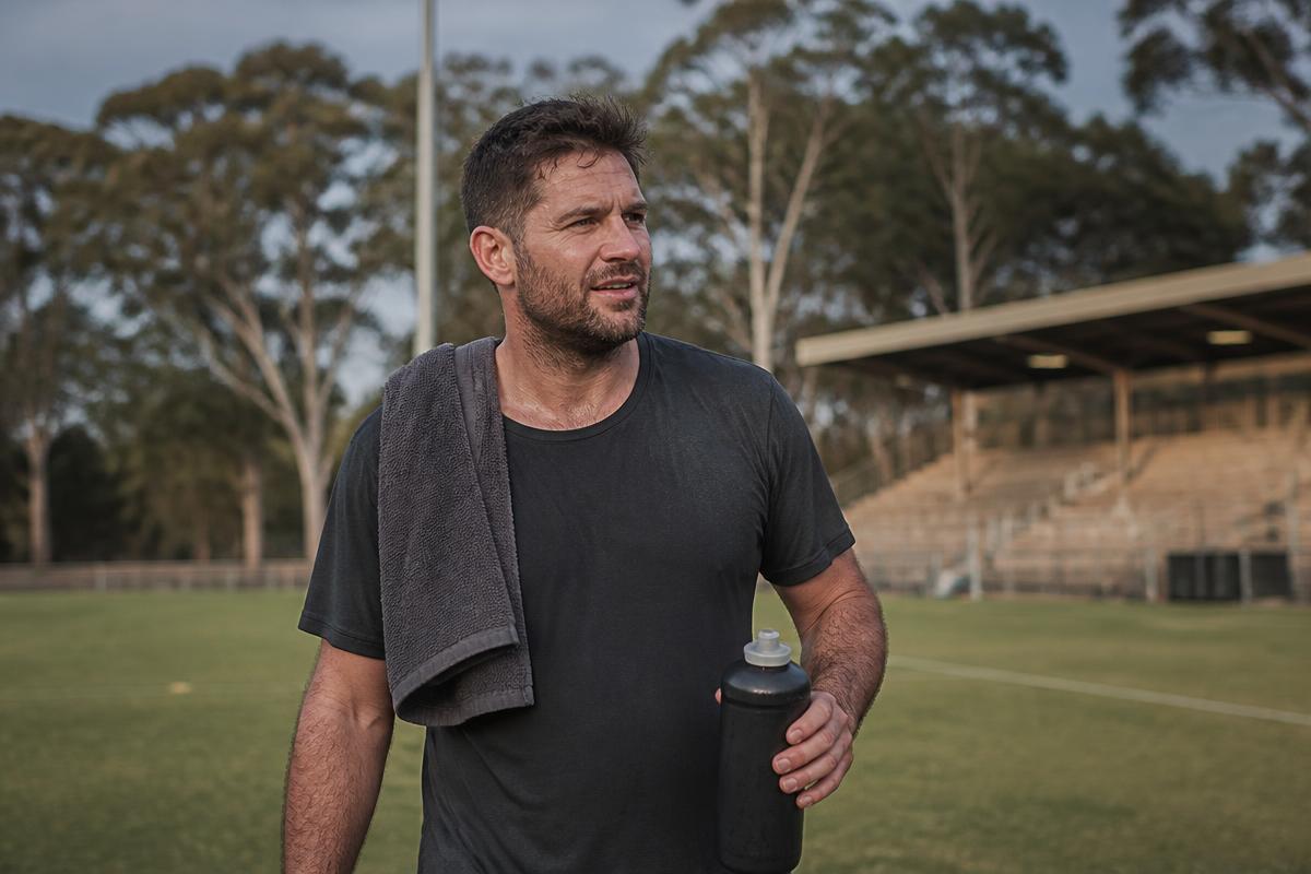 Recreational athlete standing on a community sports oval at first light, towel over one shoulder and water bottle in hand, after a training session